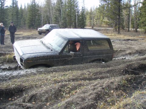 1985 Ford Bronco - A black 1980s Ford Bronco is stuck deep in mud, with another SUV visible in the background.