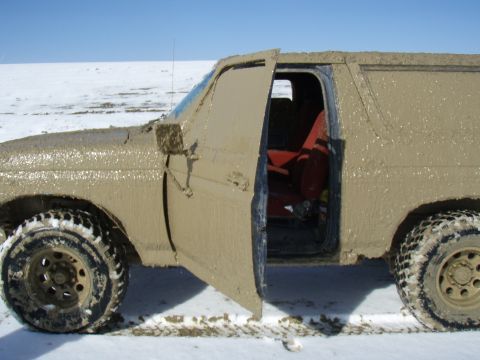 1985 Ford Bronco - This 1985 Ford Bronco is heavily covered in mud, with its driver's side door ajar, revealing a red interior.