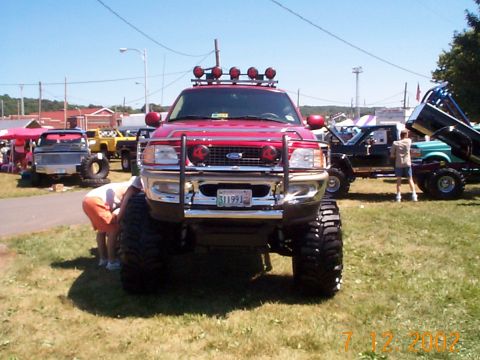 1978 Ford Bronco - A lifted red Ford truck with multiple off-road lights and a custom bumper is parked on grass at an outdoor event.