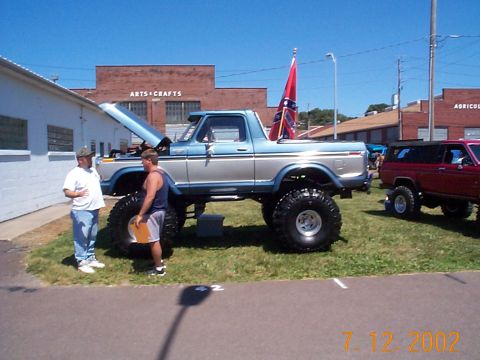 1978 Ford Bronco - Two men look over a heavily modified 1978 Ford Bronco at an outdoor event. A red SUV is visible in the background.