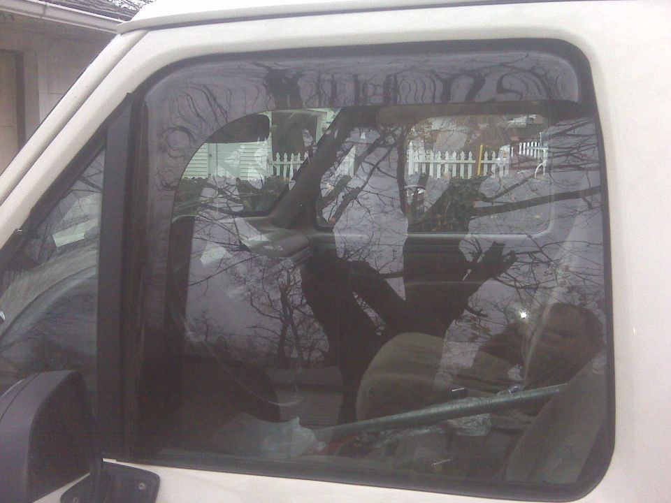 Side window of a white vehicle showing the interior and a reflection of trees and a white picket fence