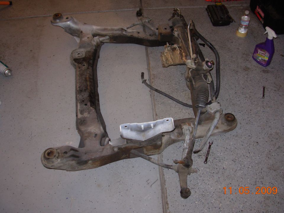 Removed front subframe with steering rack and sway bar on garage floor, cleaning supplies nearby