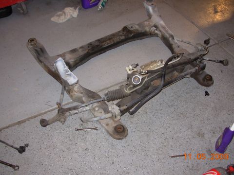 1991 Ford Taurus - A disassembled subframe and steering rack assembly lies on a concrete floor, with tools and cleaning supplies nearby.