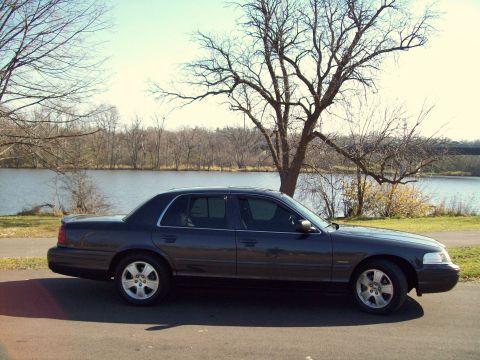 2004 Ford Crown Victoria - A dark gray sedan is parked on the side of the road next to a body of water with trees in the background.
