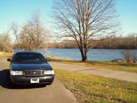 2004 Ford Crown Victoria - A dark 2004 Ford Crown Victoria is parked on a paved path next to a lake on a clear day.