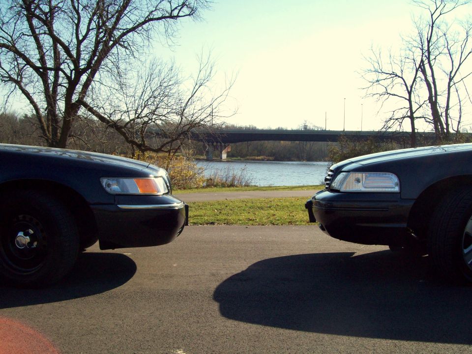 Two dark Ford sedans facing each other front-to-front by a river with a bridge behind.