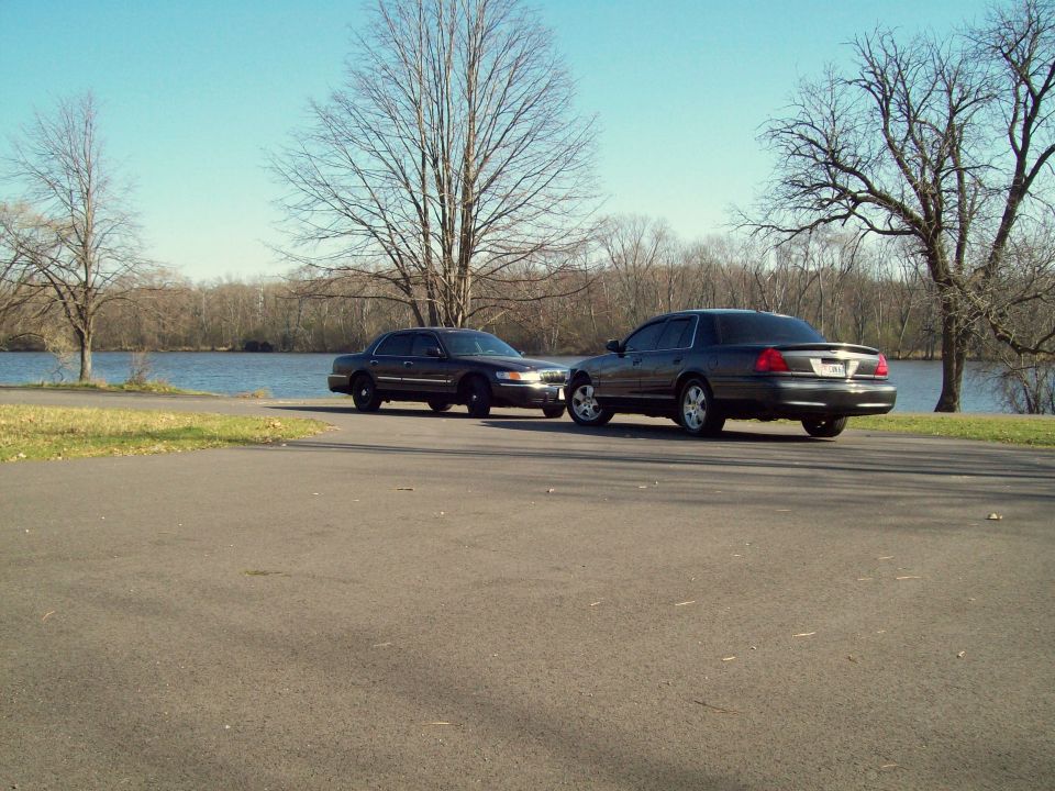 Charcoal Ford Crown Victoria and black Mercury Grand Marquis parked nose-to-nose beside a lake