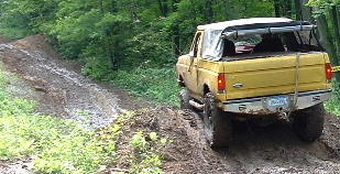 2002 Ford F-350 - Yellow pickup truck navigates a challenging muddy incline on a wooded trail.