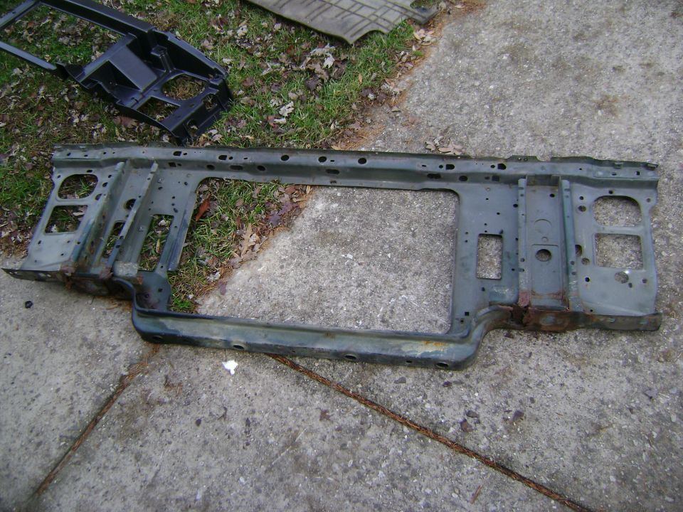 Radiator support frame for a vehicle, lying on concrete with grass in background. Another black metal piece is nearby