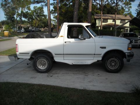 1994 Ford Bronco - This white Ford Bronco is shown from the side, parked on a concrete driveway with trees and houses in the background.