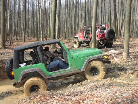1986 Ford Bronco - Two Jeeps navigate a muddy trail in a wooded area.