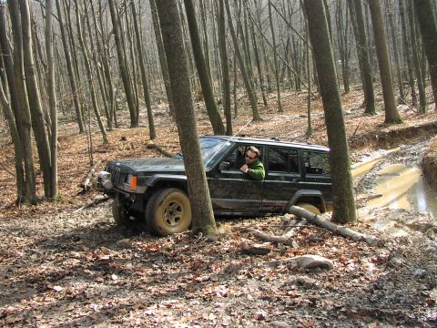 1986 Ford Bronco - A Jeep Cherokee XJ drives through a muddy, wooded area with fallen leaves on the ground.