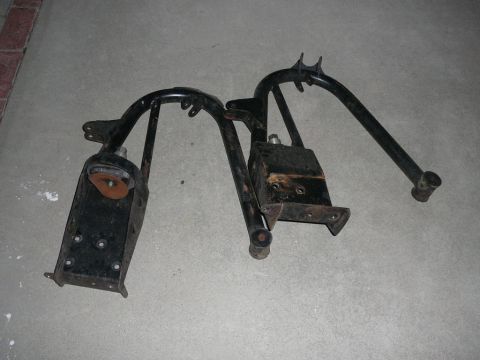 1988 Ford Bronco - Two black metal vehicle suspension components are laid out on a concrete floor, showing signs of wear and rust.