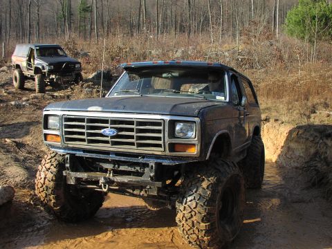 1986 Ford Bronco - Two modified off-road vehicles navigate a muddy trail in a wooded area.