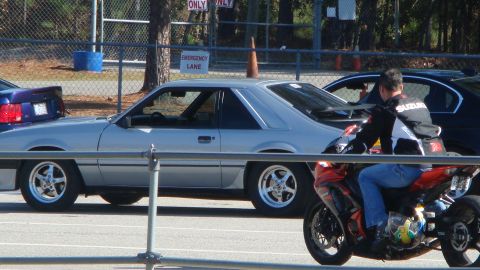 1986 Ford Mustang - A silver 1986 Ford Mustang is parked at an event, with a motorcycle and rider nearby.