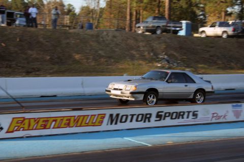 1986 Ford Mustang - A silver coupe with a hood scoop races down a track, with other vehicles and spectators in the background.