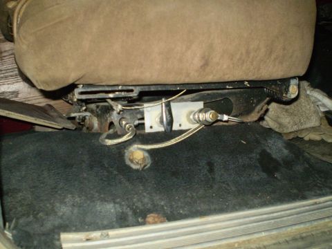 1986 Ford Bronco - The underside of a vehicle seat reveals its mechanical components and wiring, with the carpeted floor visible below.