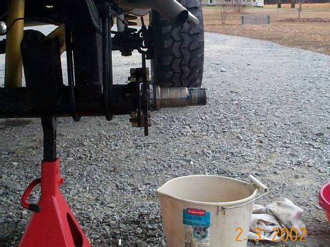 1978 Ford Bronco - Close-up of a vehicle's rear axle and brake assembly, supported by a red jack stand, with a white bucket and rag on the gravel ground.