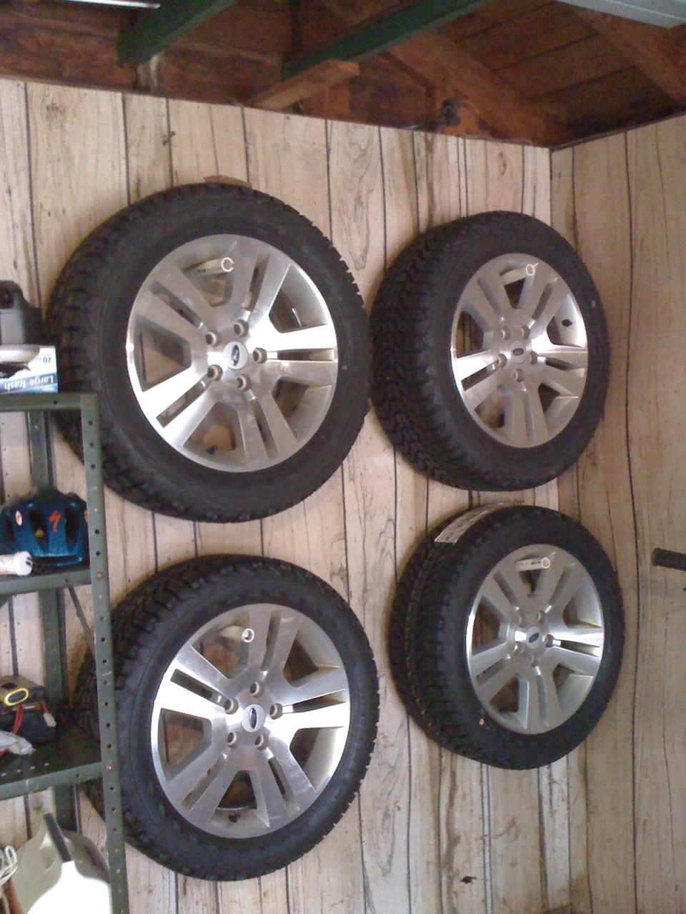 Four silver rims with tires mounted on a wood-paneled wall