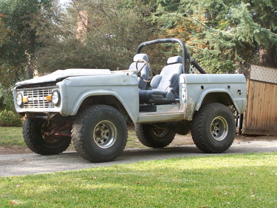 Gray Ford Bronco with roll cage and aftermarket wheels, parked on a gravel driveway