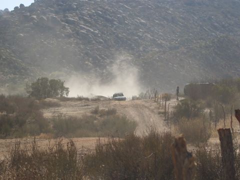 1986 Ford Bronco - A vehicle races down a dusty dirt road, kicking up a large cloud of dust against a backdrop of rocky hills.