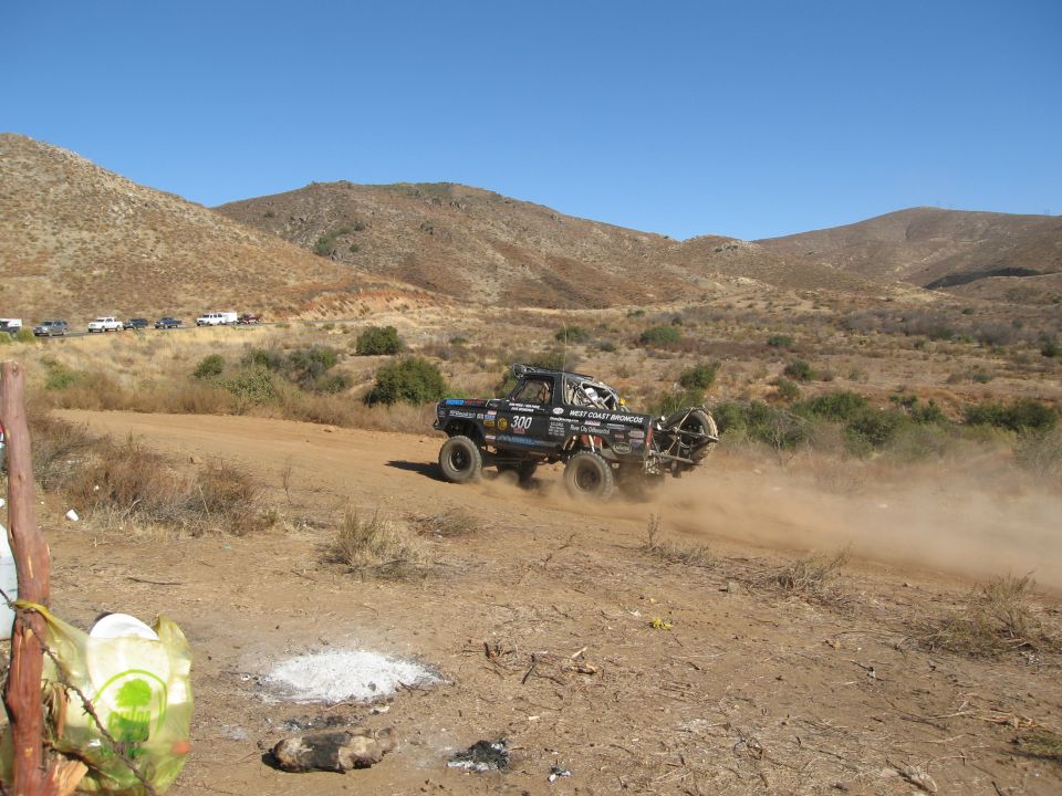 1986 Ford Bronco modified for off-road racing, kicking up dust on a dirt track. Spare tire mounted on the back