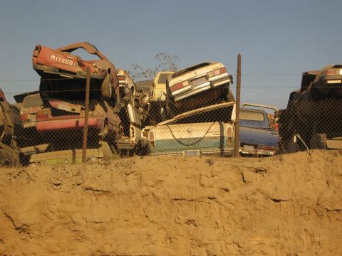 1986 Ford Bronco - Several damaged vehicles are stacked and piled up in a junkyard under a clear sky.
