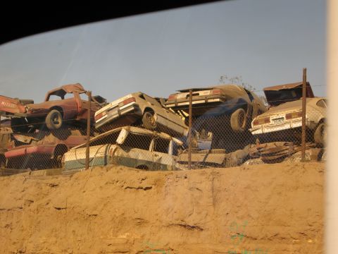 1986 Ford Bronco - A junkyard scene shows multiple wrecked vehicles piled high against a fence under a clear sky.