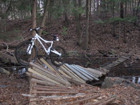 1981 Garelli Rally Sport - A white full-suspension mountain bike is positioned on a makeshift wooden bridge crossing a stream in a wooded area.