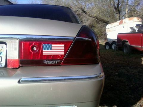 2000 Mercury Grand Marquis - A silver sedan's rear view shows a tail light adorned with an American flag sticker and SCT badging.