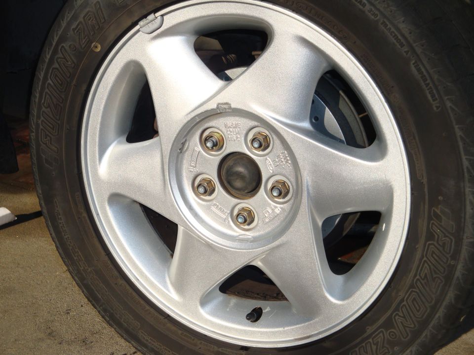 Close-up of a silver 5-spoke car wheel with a Fuzion ZRi tire. Lug nuts and part of the brake rotor are visible
