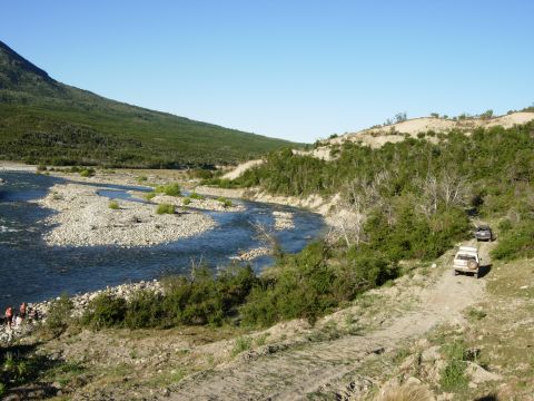 1986 Ford Bronco - Two SUVs navigate a dirt track alongside a rocky riverbed under a clear blue sky.