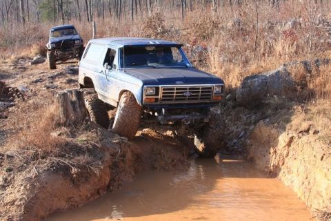 1986 Ford Bronco - Two off-road vehicles tackle a challenging muddy terrain with deep ruts and water obstacles.