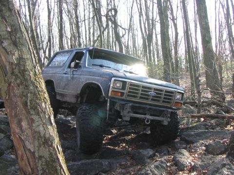 1986 Ford Bronco - A lifted Ford Bronco navigates a rocky trail through a dense forest.
