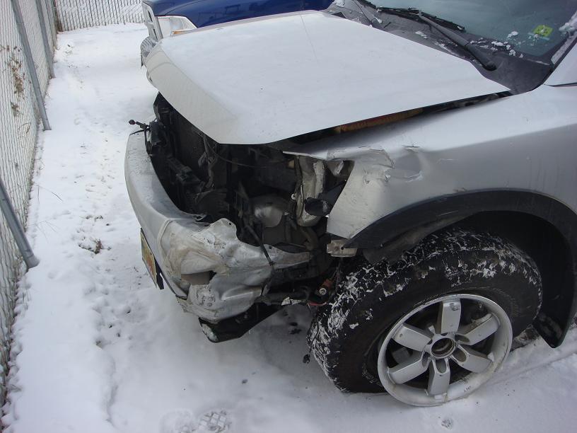 Silver pickup truck with severe front-end damage in snow. Another vehicle is visible behind it