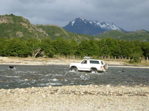 1986 Ford Bronco - A white 1986 Ford Bronco navigates a rocky riverbed with a scenic mountain backdrop.