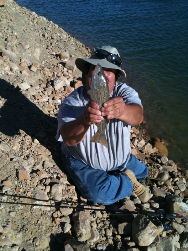Man in hat and sunglasses holds a striped fish near the water's edge with fishing rods on the rocky shore