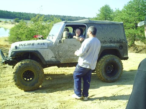 1990 Ford Bronco - A heavily modified Jeep Wrangler, covered in mud, sits on a sandy track with people around it.