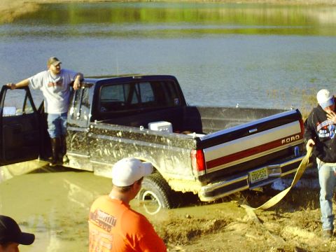 1990 Ford Bronco - A Ford F-Series truck is stuck in the mud near a body of water, with people around it.