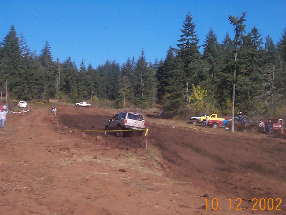 White SUV on muddy off-road course with parked trucks and pine trees behind, dated 10.12.2002