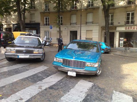 1991 Ford Crown Victoria - Two distinct vehicles, a blue Cadillac and a black Ford Crown Victoria, are parked on a city street.