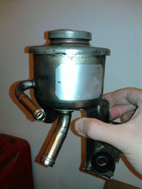 1983 Ferrari Enzo - Metal power steering fluid reservoir with hose connections and cap, held in hand.