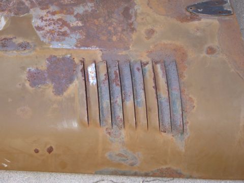 1988 Ford Bronco - Close-up of a set of stamped louver vents cut into a tan-painted sheetmetal panel, with heavy scaling rust, bubbled paint and bare primer v…