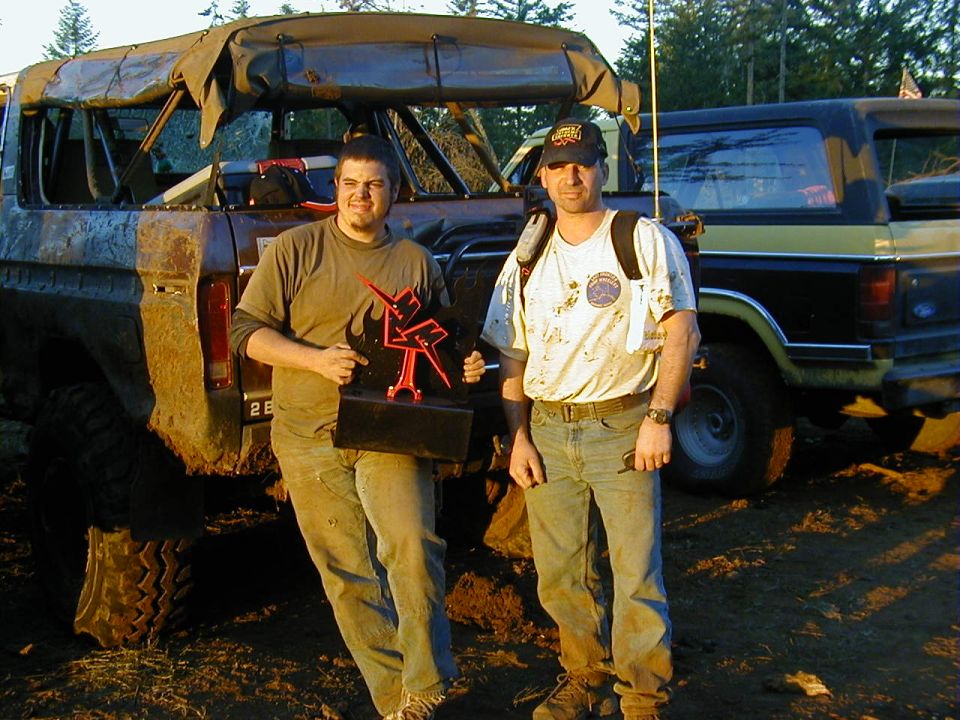 Two men holding a trophy beside muddy off-road SUVs at an outdoor wheeling event