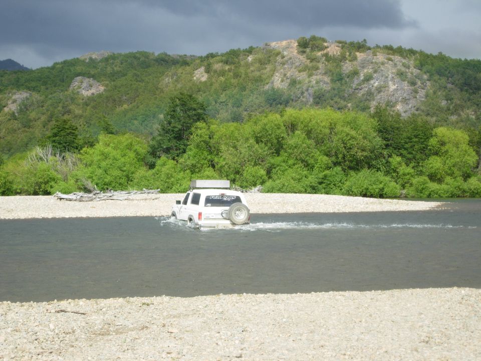 White SUV with roof box and tailgate spare fording a wide river in a forested valley