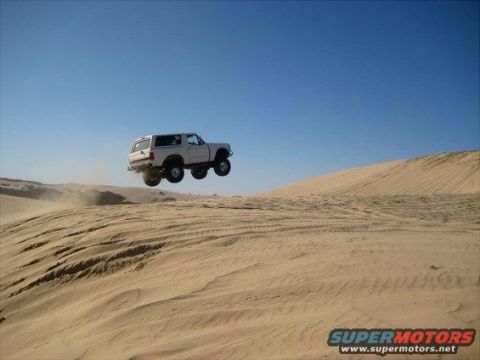 1991 Ford Bronco - A white SUV is captured mid-air, jumping over sand dunes under a clear blue sky.