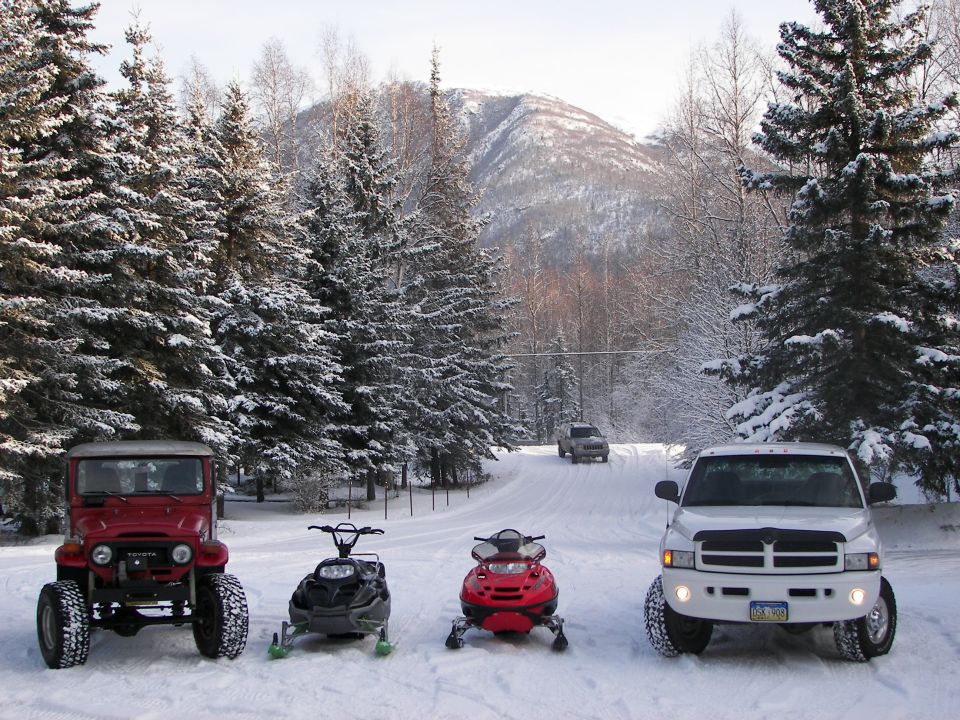Red Toyota Land Cruiser, two snowmobiles, and white Dodge Ram parked on a snowy road with mountains behind