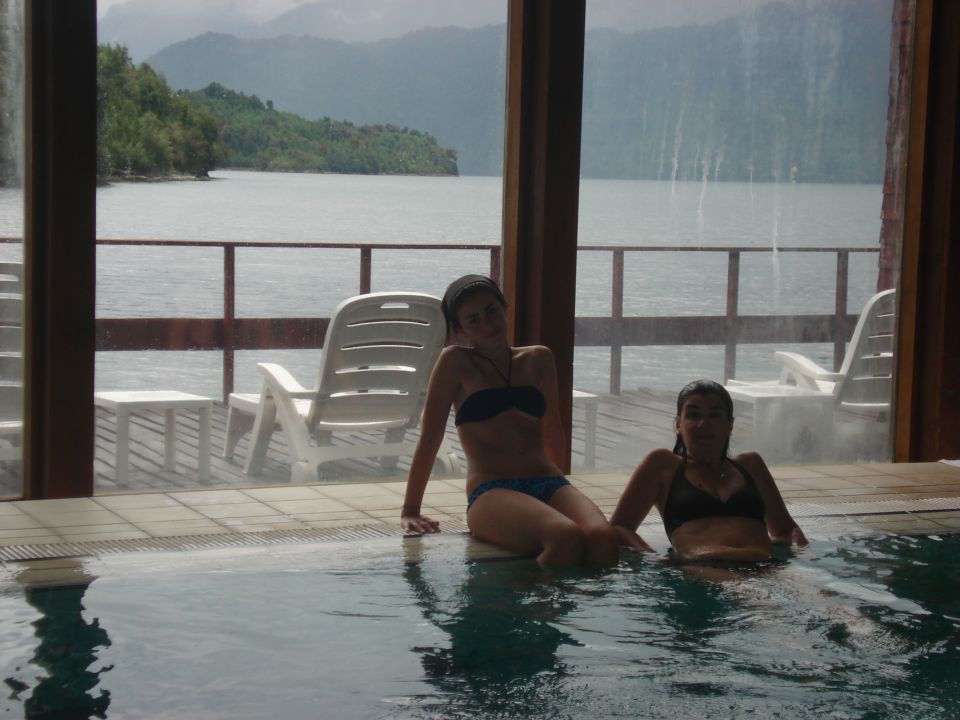 Two women relax in a pool overlooking a lake and forested mountains