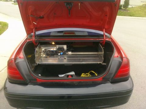 2002 Ford Crown Victoria - The open trunk of a red sedan reveals a custom metal shelf installed with various electronic components and wiring.