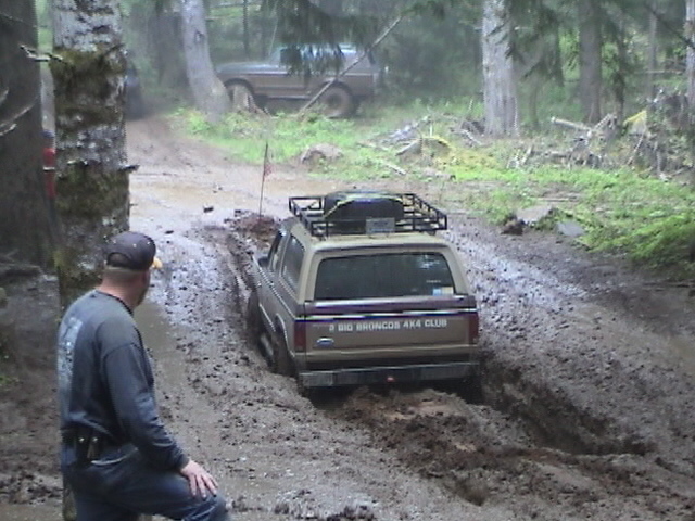 Tan Ford Bronco stuck in deep mud on forest trail while a man watches